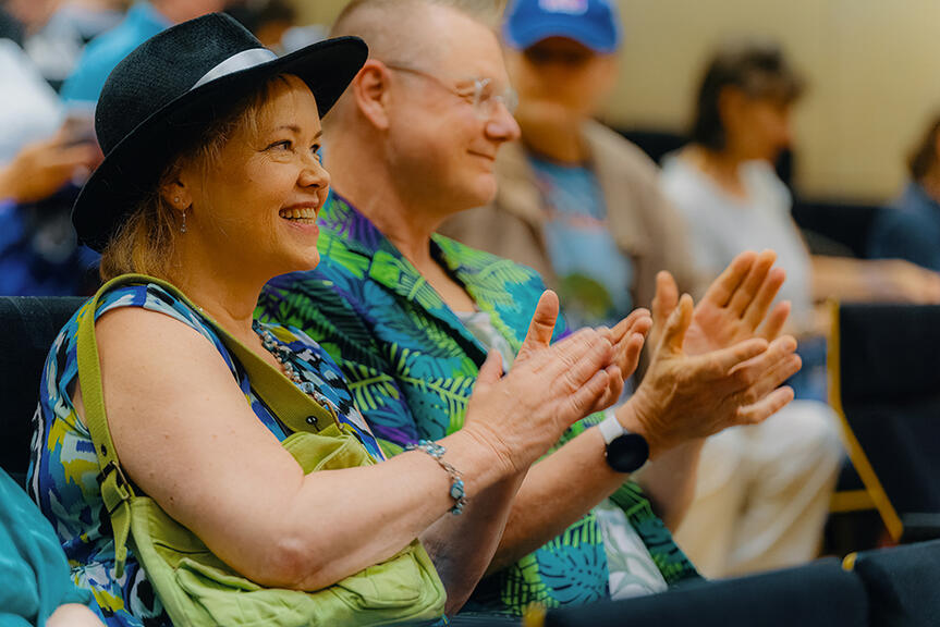 A woman and a man are smiling in the audience and clapping with their hands.