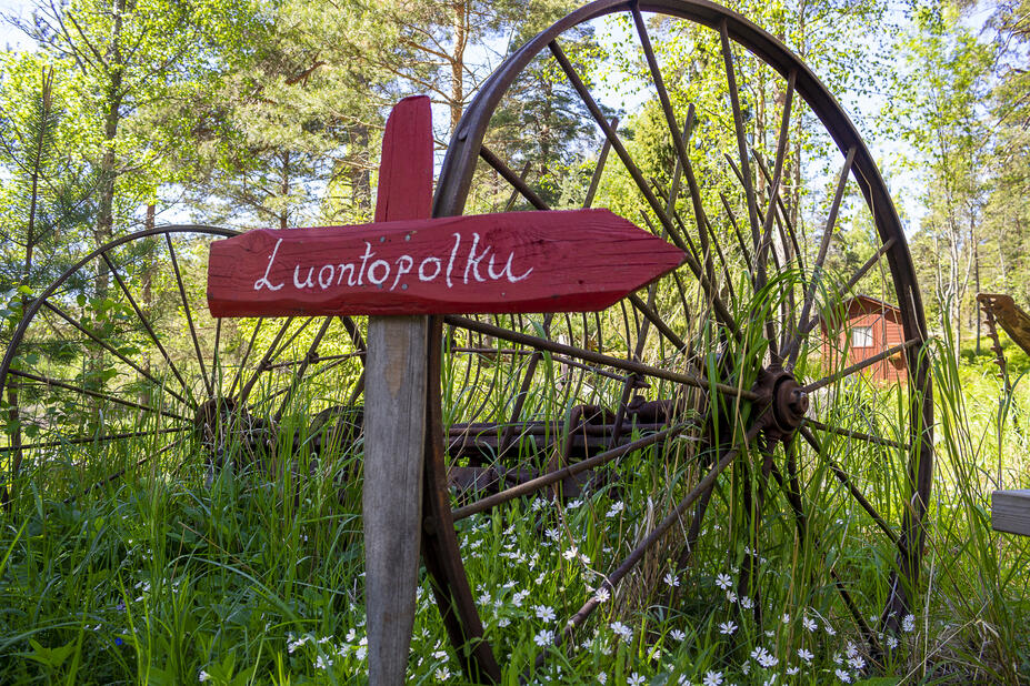 Punainen luontopolun kyltti vanhaa rattaan rautaista rengasta vasten.