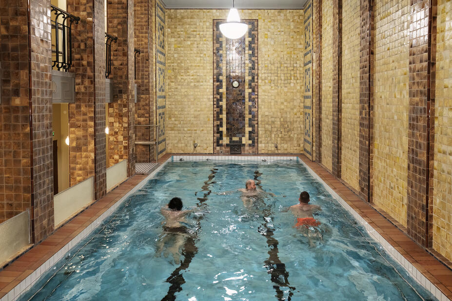  Kolme ihmistä ui Yrjönkadun uimahallin kolmannen kerroksen uima-altaassa. Seinien kaakelit ovat okran ja ruskean sävyisiä.