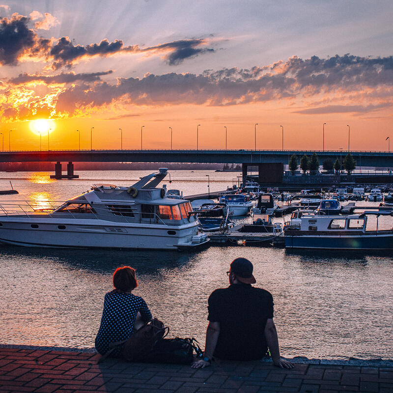 Kaksi ihmistä istuvat laiturilla ja katsovat veneitä ja auringonlaskua.