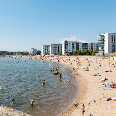 Ihmiset oleskelevat hiekkarannalla, taustalla kerrostaloja.