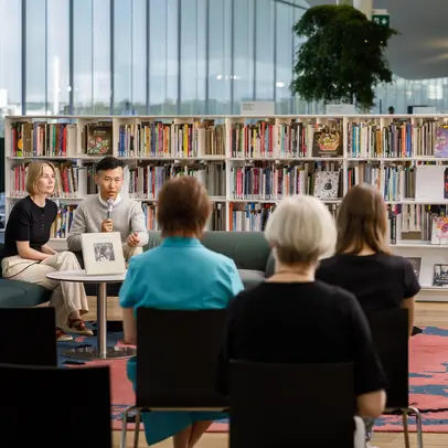 An author and an interviewer sit on a sofa at the Oodi Central Library, speaking into a microphone in front of an audience.