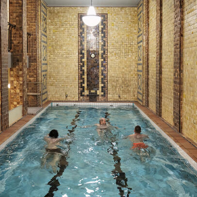  Kolme ihmistä ui Yrjönkadun uimahallin kolmannen kerroksen uima-altaassa. Seinien kaakelit ovat okran ja ruskean sävyisiä.