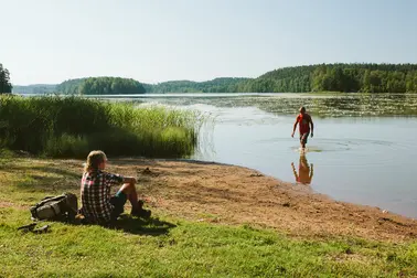 Henkilö istuu rannalla, toinen henkilö kahlaa vedesä.