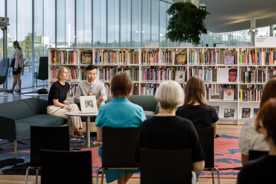 An author and an interviewer sit on a sofa at the Oodi Central Library, speaking into a microphone in front of an audience.