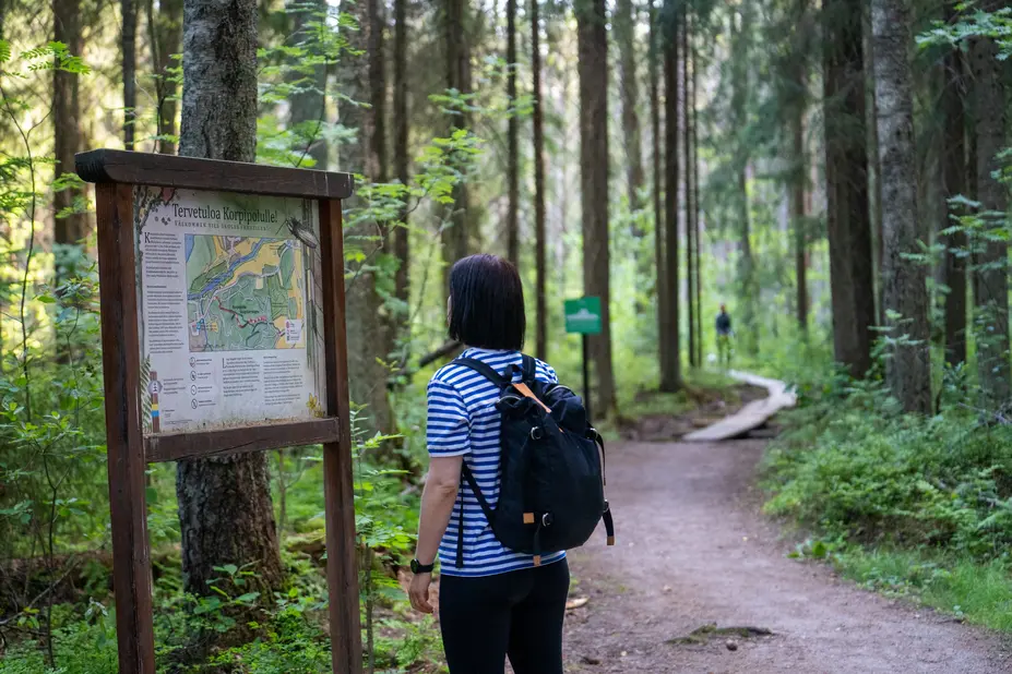 Retkeilijä tutkii luontopolun opastaulua. Taustalla metsään johtavat pitkospuut.
