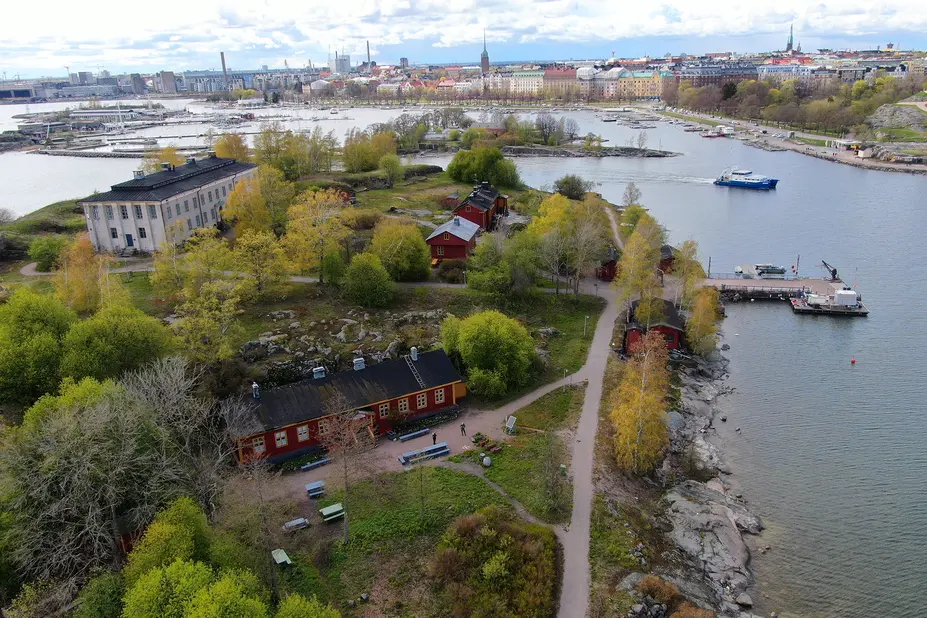 Harakan saaren dronekuvassa näkyy saaren rakennuksia ja polkuja, ympäröivä meri sekä taustalla kaupunki