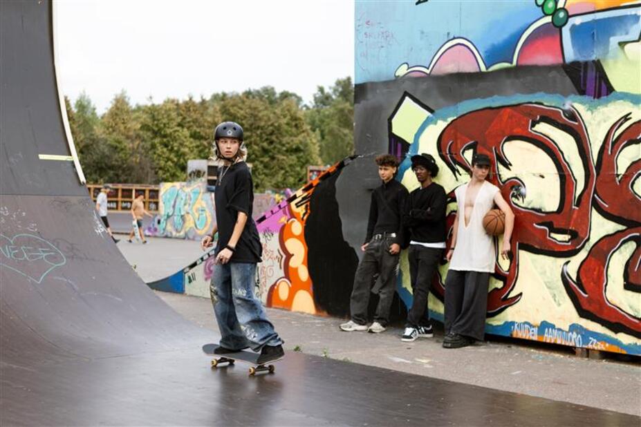 Four young people in a skate park. A young person in a black shirt and helmet is skateboarding in the foreground. Three young people, two in black tops and one in a white top, lean against a graffiti wall in the background. The young person in the white top has a basketball under their arm.
