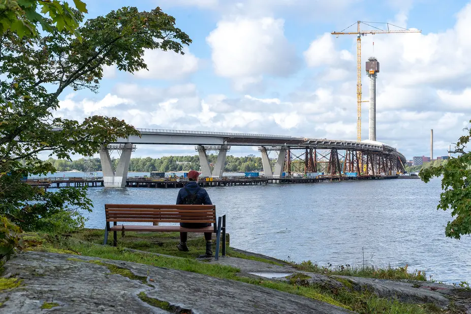 Ulkoilija istuu penkillä ja katsoo rakentuvaa Kruunuvuorensiltaa. 