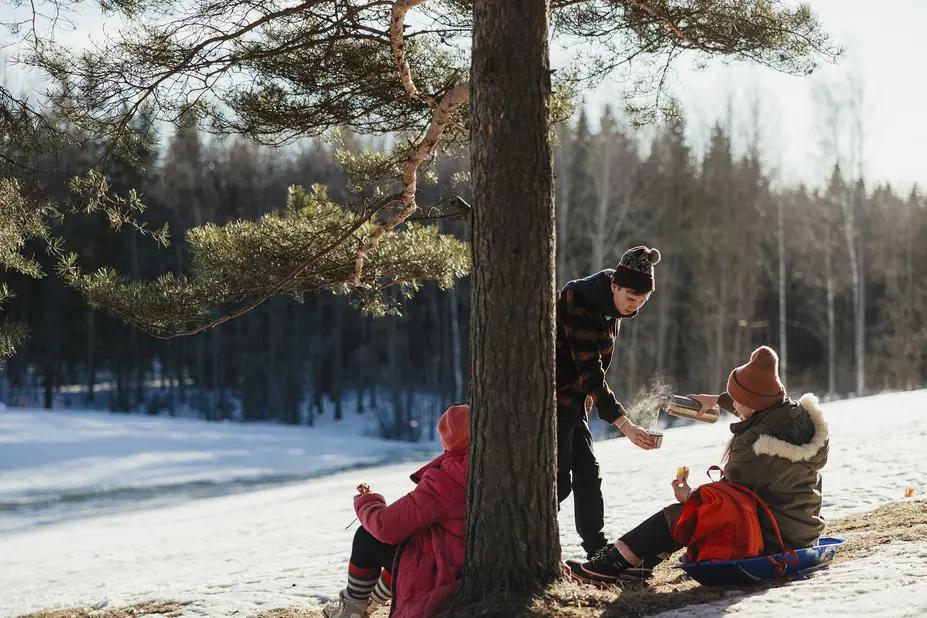 Kolme henkilöä Paloheinän ulkoilualueen rinteessä, yksi kaataa toiselle kuumaa juomaa. 