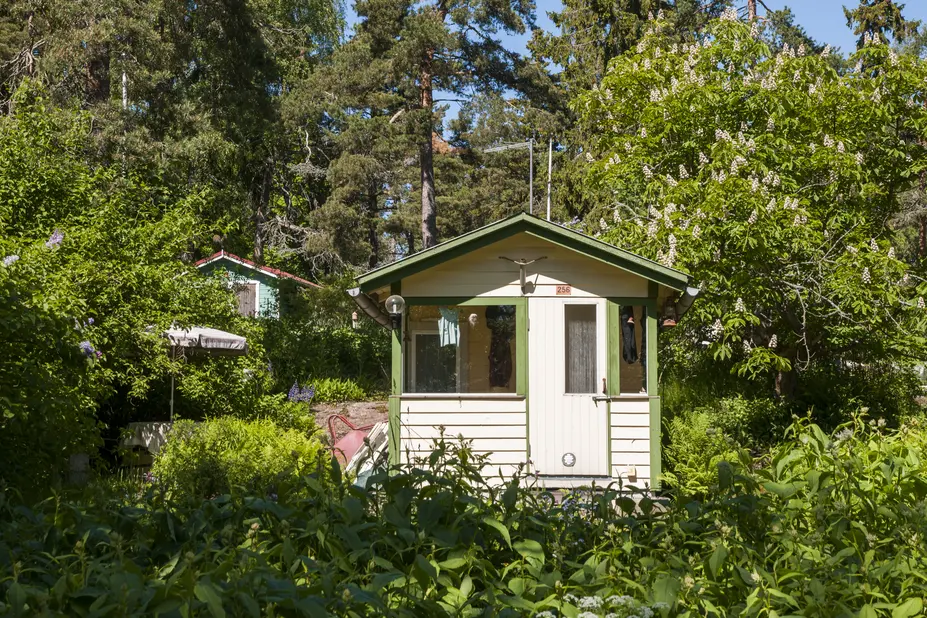 Gräddfärgad sommarstuga i ett grönskande landskap.