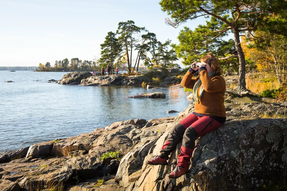 Nainen istuu kalliolla ja kiikaroi merelle