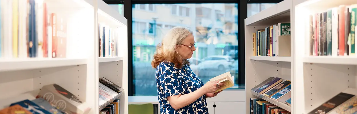 Nainen lukee kirjaa Maunulan kirjaston kirjahyllyjen välissä.
