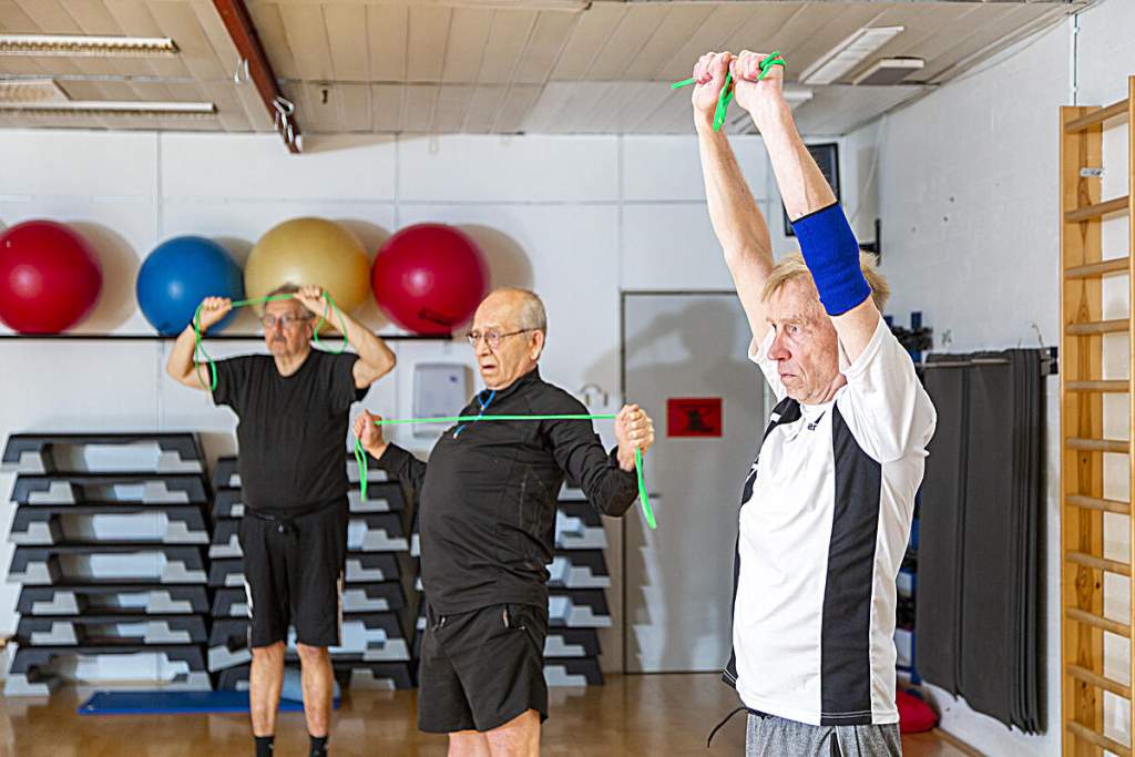 In a gym session for men, three older men are training using rubber resistance bands.