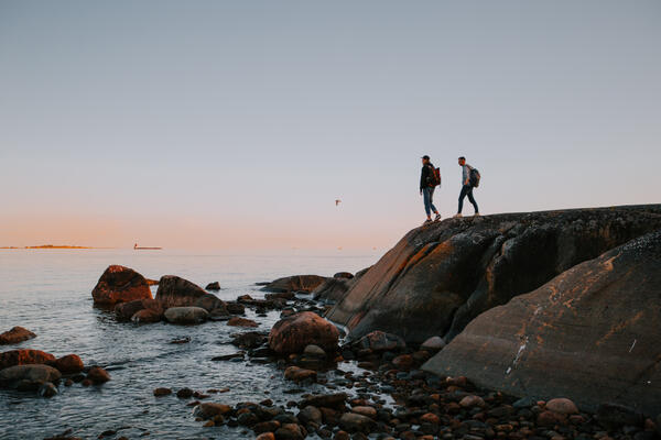 Kaksi henkilöä seisoo rantakalliolla auringon laskiessa.