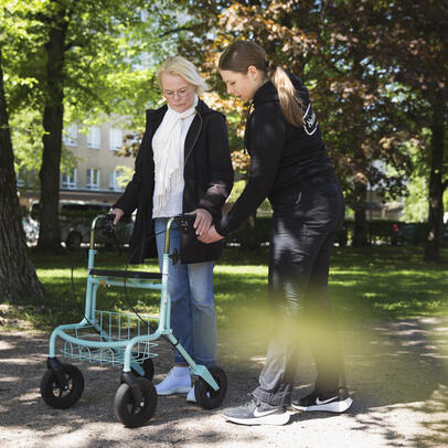 Työntekijä auttaa vanhusta rollaattorin kanssa ulkona.