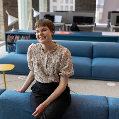 Maisema-arkkitehti Sofia Kangas nojaa hymyillen sohvaan työpaikkansa suuressa toimistohuoneessa, jossa on moderneja huonekaluja ja valaisimia. 