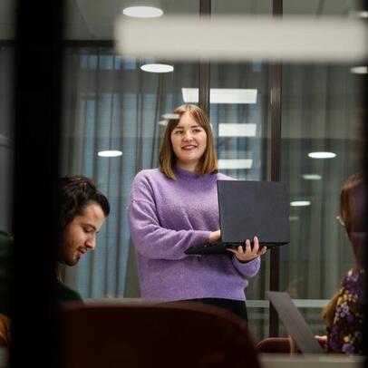 Nainen seisoo läppäri kädessään ja hymyilee.