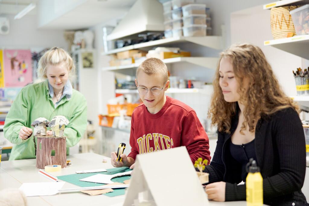 Kolme opiskelijaa rakentaa kuvataiteen töitä valoisessa luokassa.