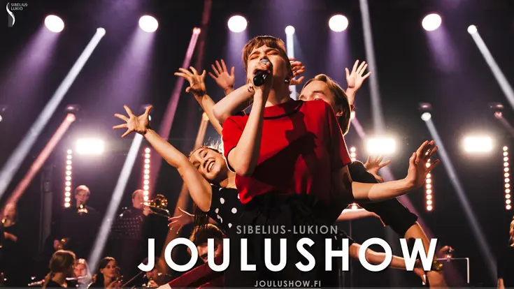 Joulushown laulaja punaisessa asussa etualalla ja tanssijoita tanssijoita kädet ylhäällä.
