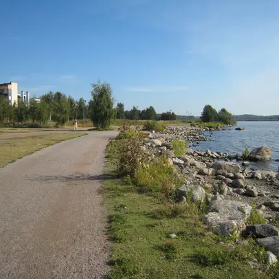 Rannassa kulkee hiekkatie ja taaempana näkyy puita ja rakennuksia.