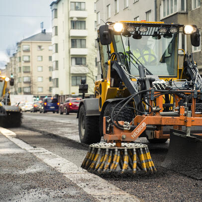 Kaksi työkonetta puhdistaa katua hiekasta, taustalla näkyy kerrostaloja.