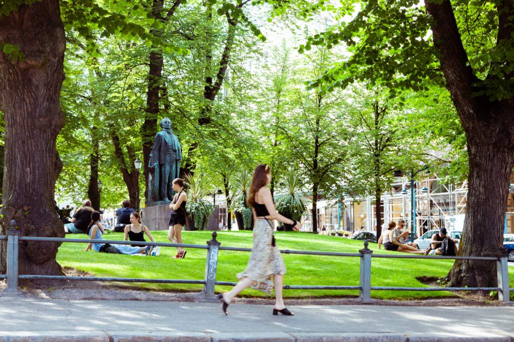Nainen kävelee kesäisellä kadulla ja ihmisiä istuskelee puiston nurmikolla.