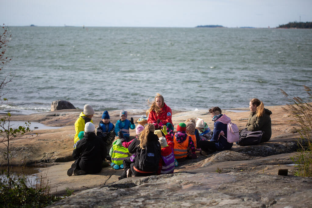Päiväkotiryhmä istuu meren äärellä kallioilla ja seuraa ympäristökasvattajaa