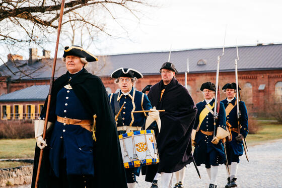 Viisi henkilöä kulkee tummissa historiallisissa asuissa Suomenlinnassa mukulakivikaduilla.