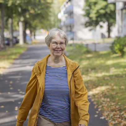 Hymyilevä seniori kävelee jalkakäytävällä puiden keskeltä.