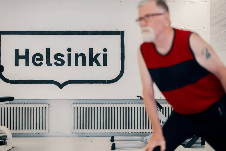 Mies liikuntasalissa urheilemassa takaseinässä maalattuna iso Helsinki kehystunnus.