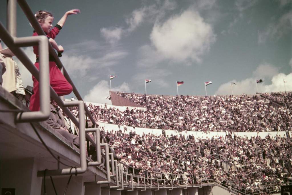 Tyttö punaisessa asussa vilkuttaa kädellään urheilijoille Olympiastadionin katsomossa vanhassa kuvassa.