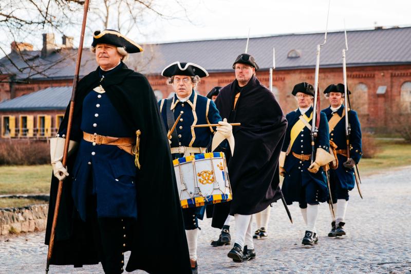 Five men in dark historical costumes walk in the streets of Suomenlinna.