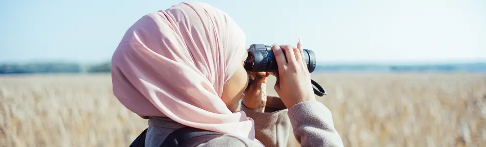 A person looking at a field landscape with binoculars, the person has a pink scarf on their head.