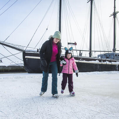 Mom and a small kid skating by the docks of old wooden boats.